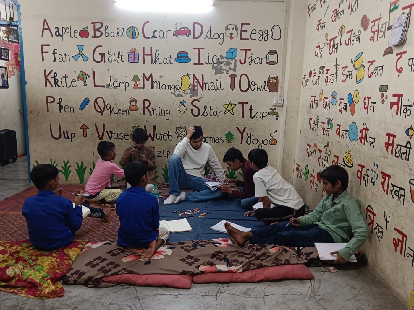 Children sitting on mats in a small group, learning in front of a hand-painted English and Hindi alphabet wall