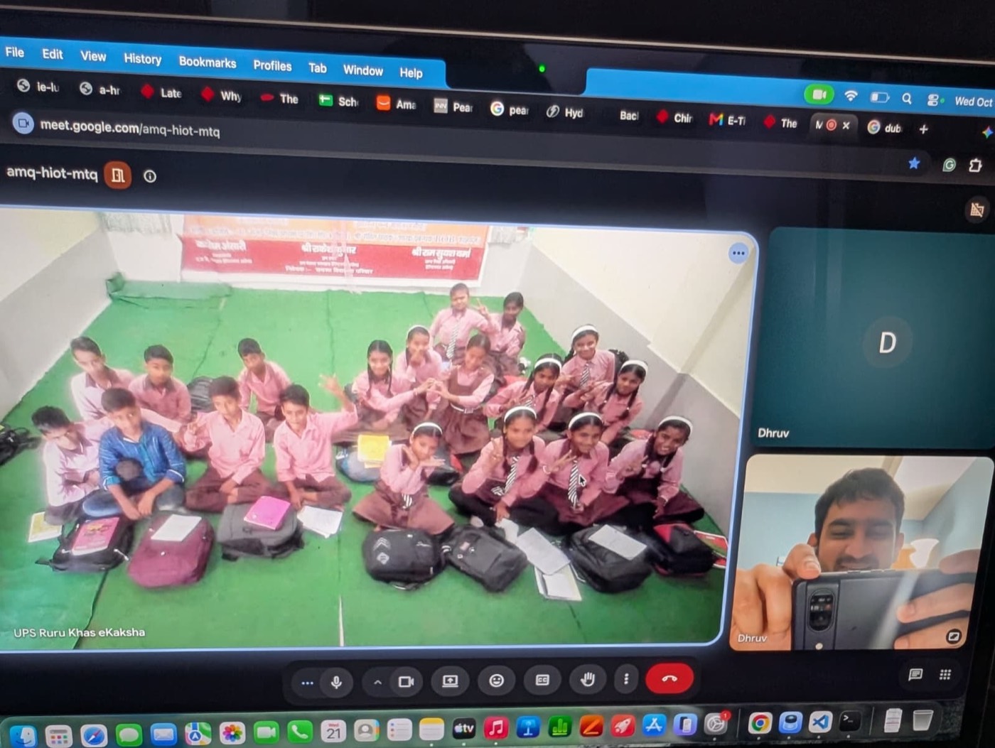 A laptop showing a virtual class: a classroom of students seated on the floor, with a volunteer educator joining by video